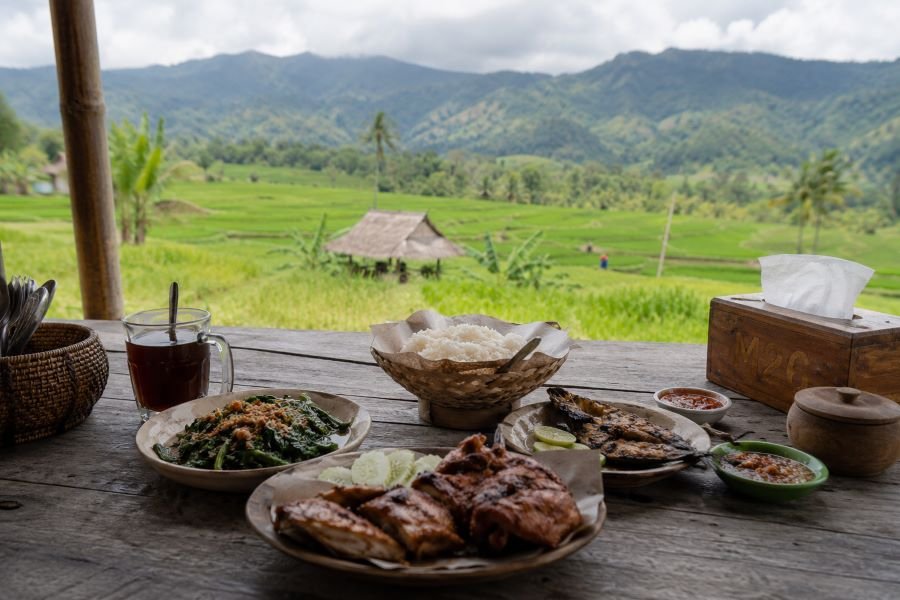 One thing people rarely explain properly about village food in Lombok is this: freshness changes everything.
Vegetables taste brighter. Chili feels sharper. Even plain rice somehow tastes… warmer? Hard to explain.
At a Tetebatu warung, meals are often cooked only after you order them. You wait a little longer, yes, but nobody really complains because the surroundings make waiting enjoyable. You stare at the rice fields. Maybe sip black coffee. Maybe hear distant roosters screaming for no reason.
Then the food arrives.
Plecing kangkung. Ayam taliwang. Fresh grilled fish. Sambal that wakes up your entire nervous system.
And somehow you keep eating even when you’re already full.